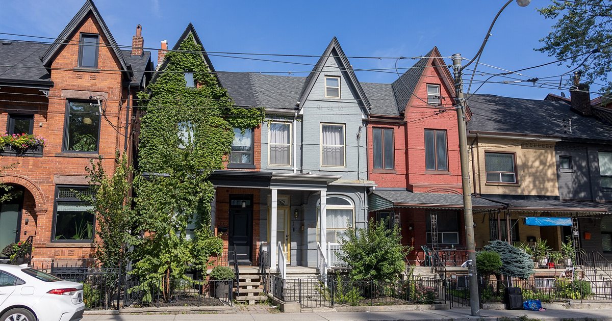 A row of brick semi-detached houses with gabled roofs and front porches on a tree-lined street in Toronto