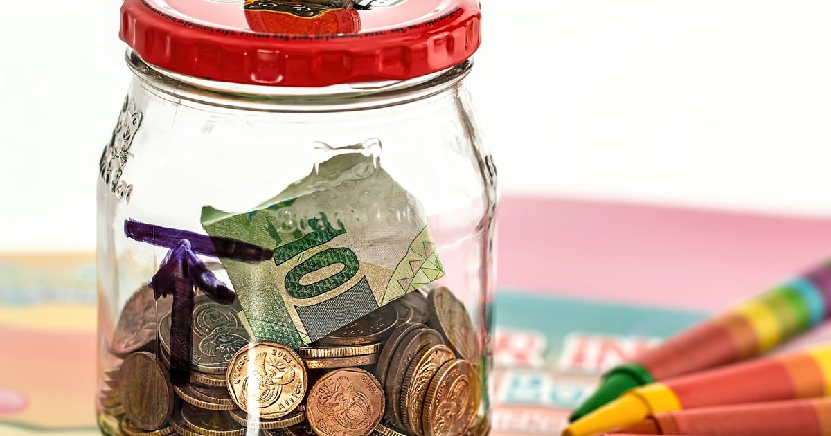 A glass jar filled with coins and folded dollar bills next to colorful crayons