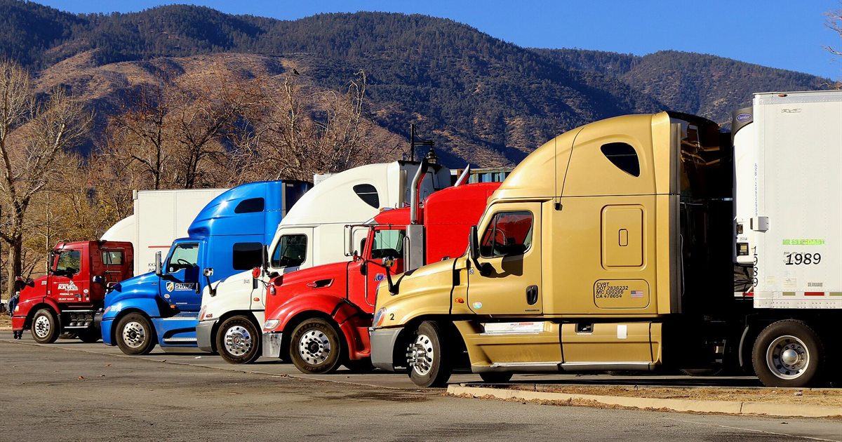 A row of brightly coloured semi-trucks parked side by side at a truck stop with mountains behind them