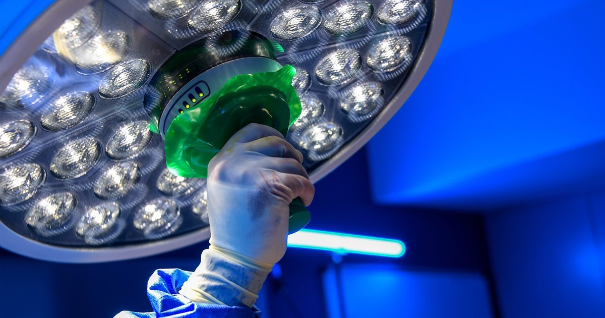 A surgeon's gloved hand adjusting a large round surgical light in a blue-lit operating room