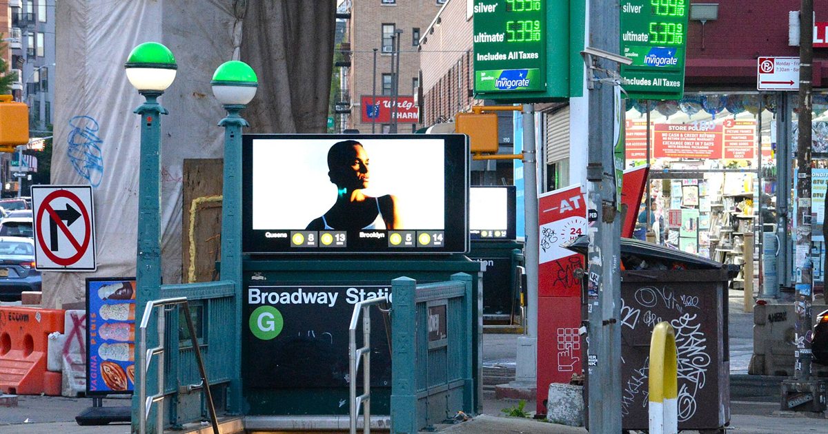 A New York street corner with a subway entrance and a gas station price sign in the background